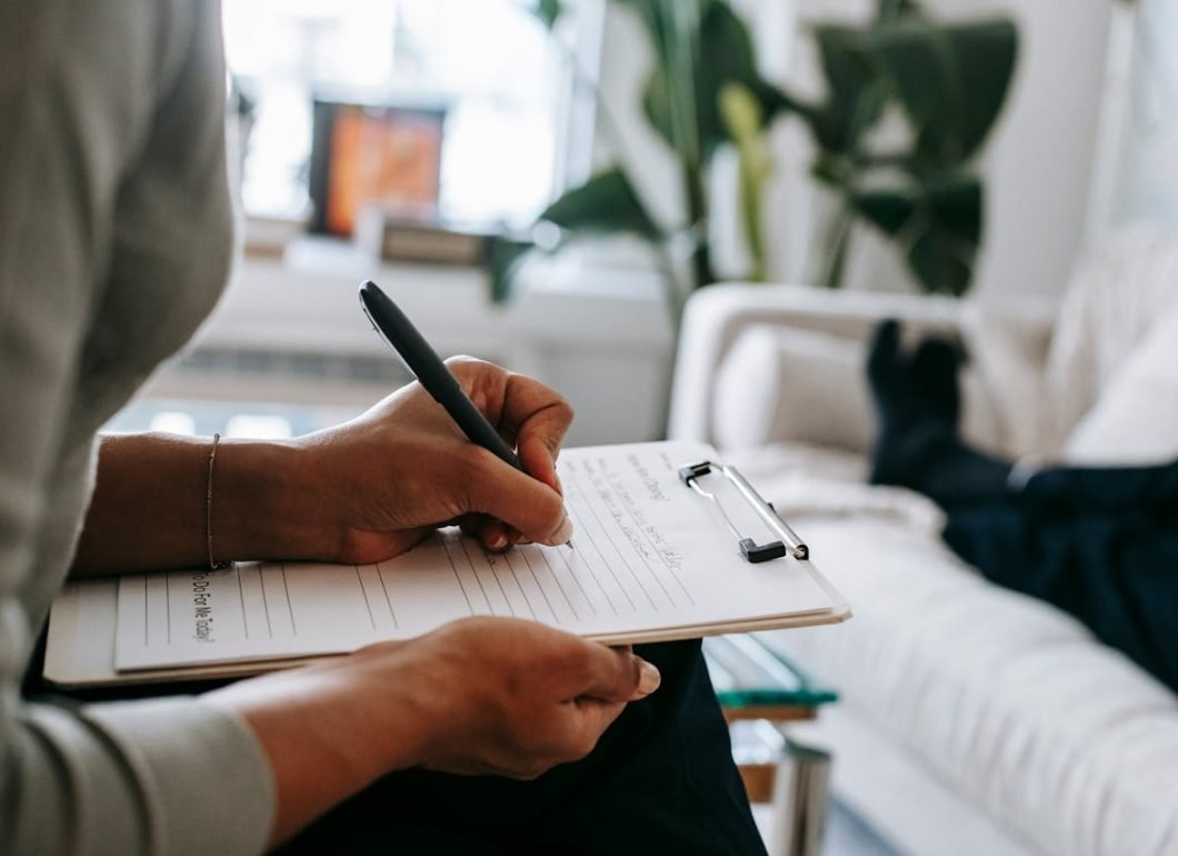 pexels-photo-5699456 Unrecognizable ethnic female therapist taking notes on clipboard while filling out form during psychological appointment with anonymous client lying on blurred background