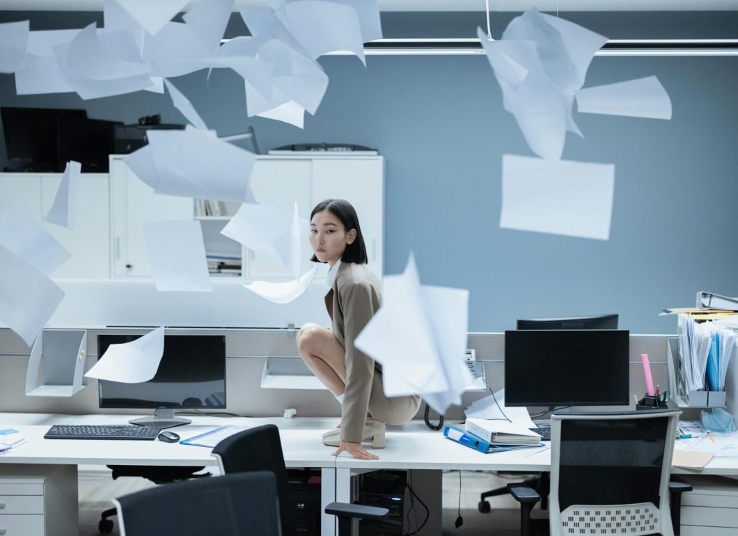 pexels-photo-8468818-8468818 Woman perched on a desk in a chaotic office with papers flying, representing workplace stress.