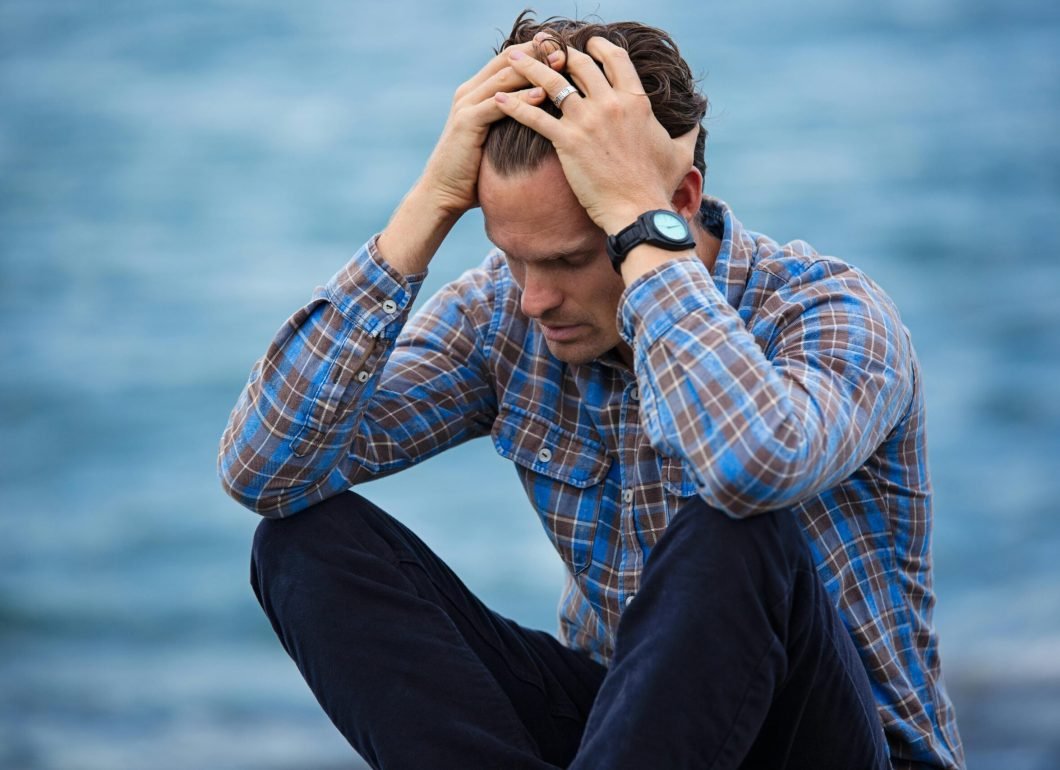 pexels-photo-897817-897817 A man in a plaid shirt sits by the water looking distressed, symbolizing stress.